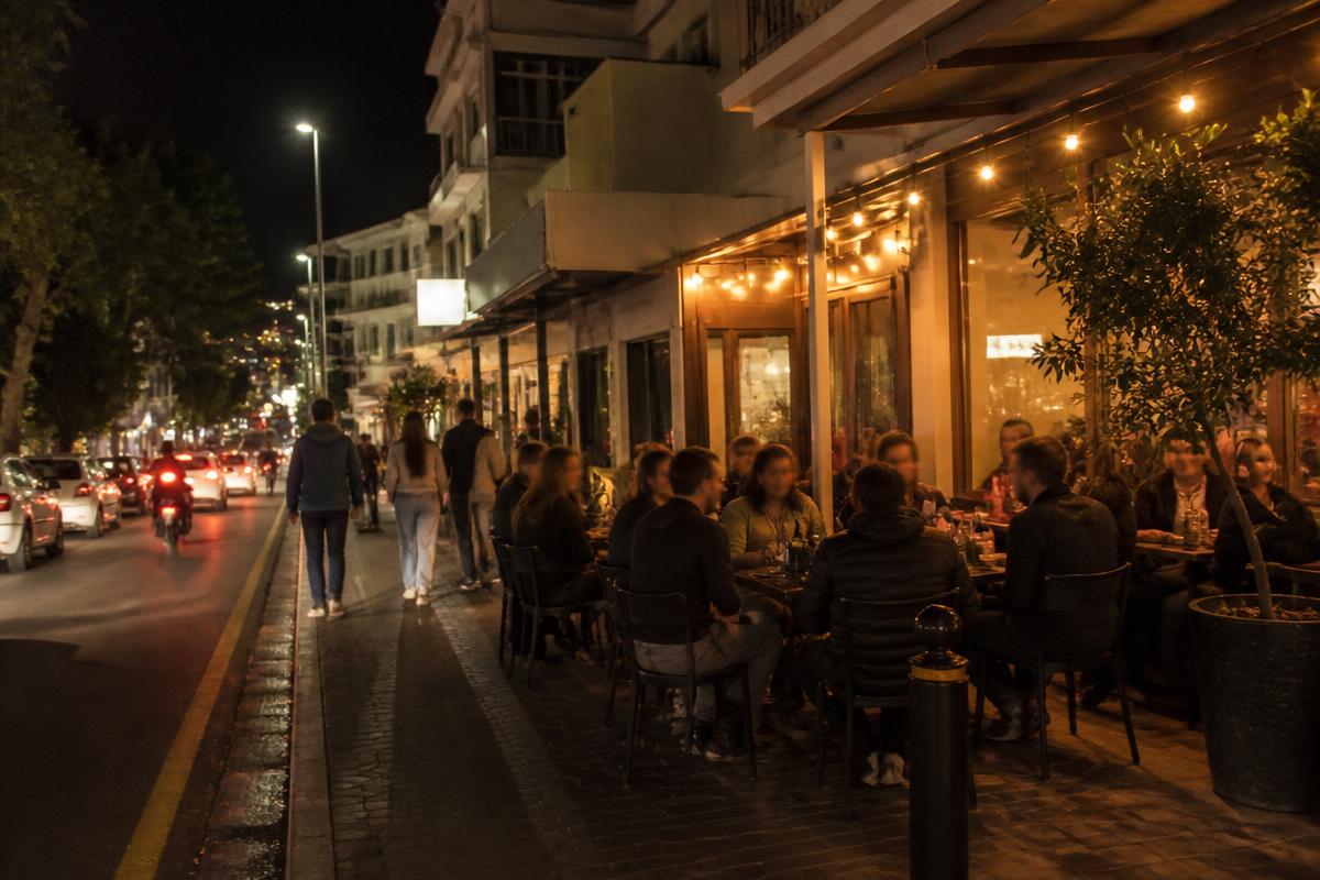 Diners on the pavement outside restaurants on a busy lit street at night